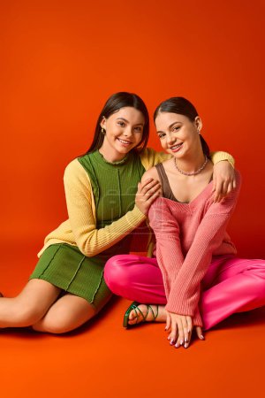 Foto de Dos adolescentes morenas con atuendo casual posando alegremente sobre un fondo naranja en un ambiente de estudio. - Imagen libre de derechos