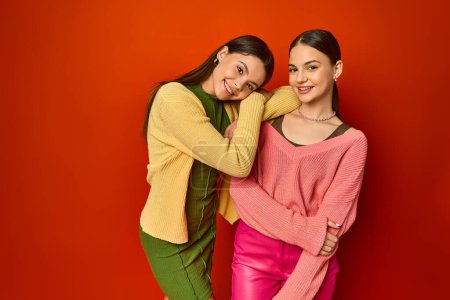 Two pretty brunettes, teenage friends, standing together in front of a vibrant red wall in a studio setting.