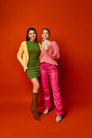 Zwei hübsche Teenager-Brünetten in lässiger Kleidung stehen zusammen vor einem leuchtend roten Hintergrund.
