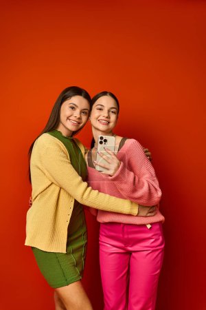 Foto de Dos guapas adolescentes, morenas y vestidas casualmente, de pie lado a lado frente a una vibrante pared roja. - Imagen libre de derechos