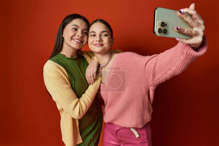 Foto de Dos bonitos amigos adolescentes capturando un momento con un teléfono celular en un estudio sobre un fondo naranja. - Imagen libre de derechos