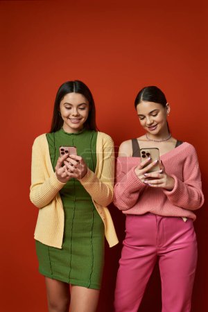Foto de Dos guapas adolescentes morenas vestidas de manera casual, paradas una al lado de la otra, sosteniendo teléfonos celulares y sonriendo. - Imagen libre de derechos