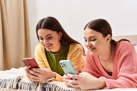 Foto de Dos mujeres jóvenes con ropa casual yacían una al lado de la otra en una cama, absortas en sus teléfonos celulares. - Imagen libre de derechos