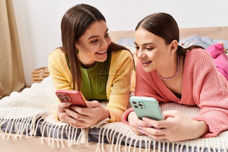 Téléchargez les photos : Deux jeunes femmes en tenue décontractée se prélassant sur un lit, absorbées dans leurs téléphones cellulaires. - en image libre de droit