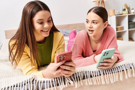 Photo for Two pretty teenage girls in casual attire, laying on a bed, engrossed in a cellphone screen together. - Royalty Free Image