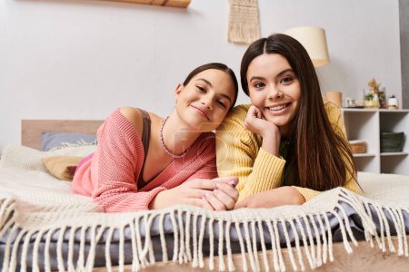 Foto de Dos amigas, guapas adolescentes vestidas de manera casual, tumbadas en una cama, sonriendo a la cámara en un momento sereno. - Imagen libre de derechos