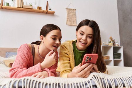 Téléchargez les photos : Deux femmes en tenue décontractée, sur un lit, souriantes, regardant ensemble un écran de téléphone portable. - en image libre de droit