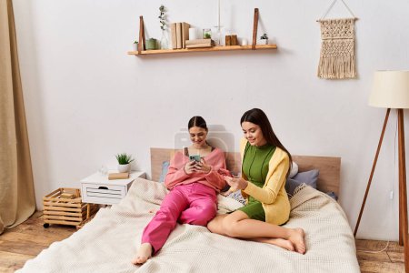 Téléchargez les photos : Deux jeunes femmes en tenue décontractée assises sur un lit, englouties dans un téléphone portable. - en image libre de droit
