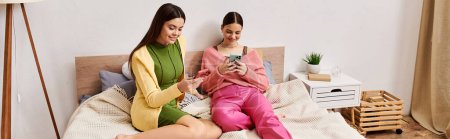 Téléchargez les photos : Deux jeunes femmes en tenue décontractée assises sur un lit, absorbées dans un téléphone portable. - en image libre de droit