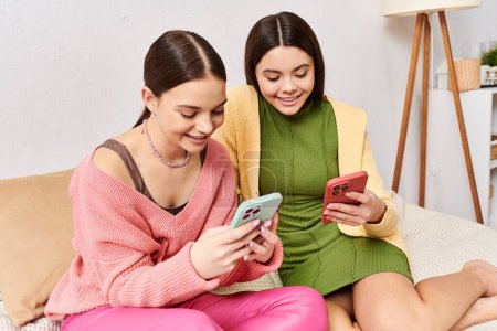 Photo for Two young women, friends, sitting on a couch engrossed in their cell phones, unaware of the world around them. - Royalty Free Image