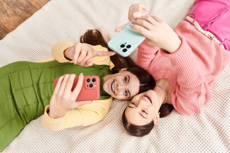 Two pretty teenage girls in casual attire, laying on a bed, taking selfies with their cell phones.