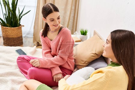 Two pretty teenage girls in casual attire sit on a bed, engaged in conversation and sharing secrets with each other.