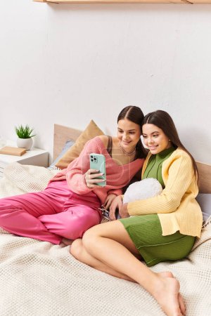 Téléchargez les photos : Deux femmes, jolies amies adolescentes, assises sur un lit et englouties dans un écran de téléphone portable. - en image libre de droit