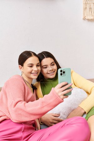 Foto de Dos adolescentes guapas vestidas con atuendo casual sentadas en un sofá, capturando un momento divertido tomando una selfie juntas. - Imagen libre de derechos