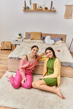 Two pretty teenage girls in casual attire sitting on the floor in front of a bed, chatting and relaxing together at home.