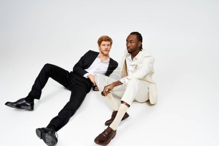 Téléchargez les photos : Deux beaux hommes multiculturels en tenue élégante assis les uns à côté des autres sur un fond blanc. - en image libre de droit