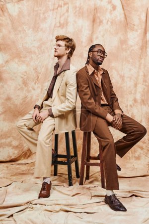 Two diverse men with dapper style sitting on top of a wooden stool.