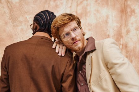 Two multicultural men dressed in dapper style hug in front of a wall.