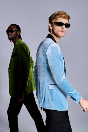 Two diverse men in dapper outfits walk confidently down an urban street wearing sunglasses.