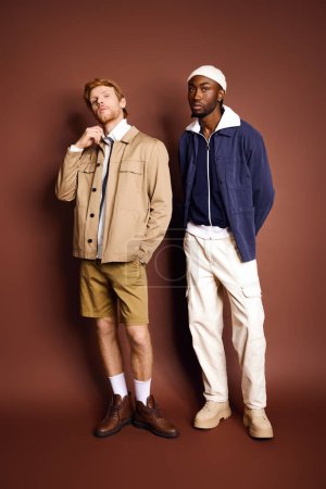 Two multicultural men with stylish attire standing next to each other in front of a brown wall.