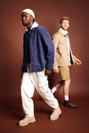 Zwei junge Männer in stylischer Kleidung gehen vor braunem Hintergrund die Straße entlang.
