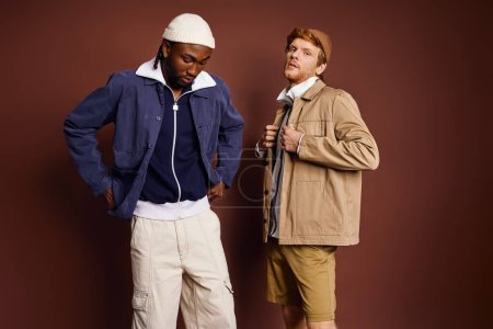 Dos hombres elegantes con antecedentes multiculturales se paran juntos con confianza frente a una pared marrón.