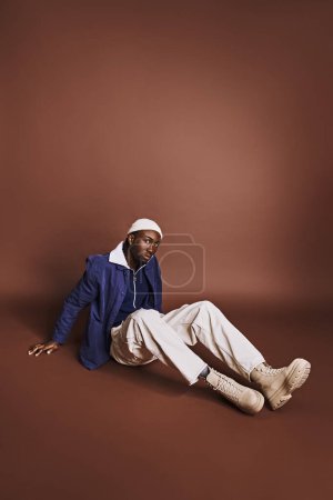 Foto de Hombre afroamericano guapo sentado en el suelo con chaqueta azul y pantalones blancos. - Imagen libre de derechos