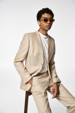 Handsome African American man in stylish sunglasses, sitting on stool in tan suit.