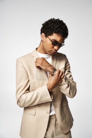 Hombre afroamericano guapo con un traje bronceado y gafas de sol elegantes ajustando su reloj.