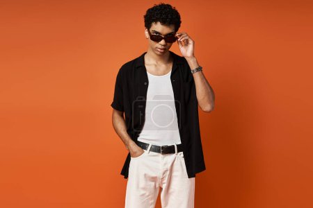 Foto de Hombre guapo con gafas de sol en camisa negra y pantalones blancos posando sobre un vibrante fondo naranja. - Imagen libre de derechos