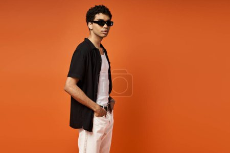 Foto de Hombre afroamericano guapo en camisa negra y pantalones blancos se levanta contra la vibrante pared naranja. - Imagen libre de derechos
