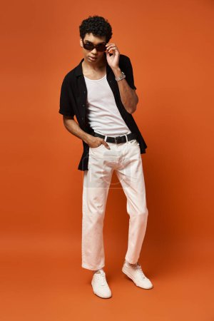 Photo for Handsome man in white pants and black shirt poses on vibrant orange backdrop. - Royalty Free Image