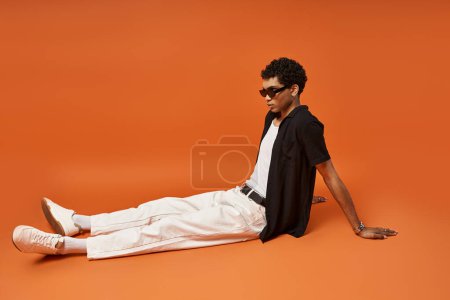 Foto de Hombre afroamericano guapo con gafas de sol elegantes acostado sobre un fondo naranja audaz. - Imagen libre de derechos
