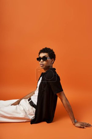 Handsome man in sunglasses and white pants relaxing on orange background.