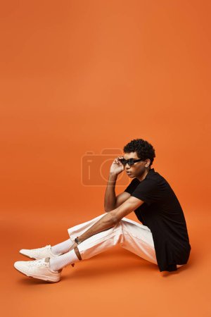 Schöner afroamerikanischer Mann sitzt mit Sonnenbrille und weißen Schuhen auf dem Boden.