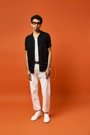 Hombre afroamericano guapo en camisa negra y pantalones blancos se para con confianza delante de un fondo naranja vibrante.