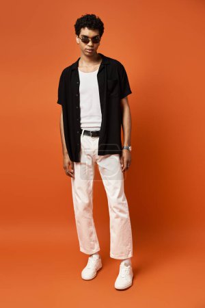 Photo for African American man in white pants and black shirt stands against bold orange backdrop. - Royalty Free Image