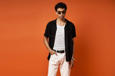 Téléchargez les photos : Bel homme afro-américain en pantalon blanc et chemise noire debout en toute confiance sur un fond orange audacieux. - en image libre de droit