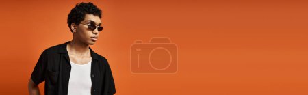 Téléchargez les photos : Un homme afro-américain à la mode se tient avec confiance dans des lunettes de soleil sur un fond orange vif. - en image libre de droit