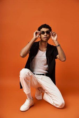 Handsome man in white pants and sunglasses crouching on orange background.