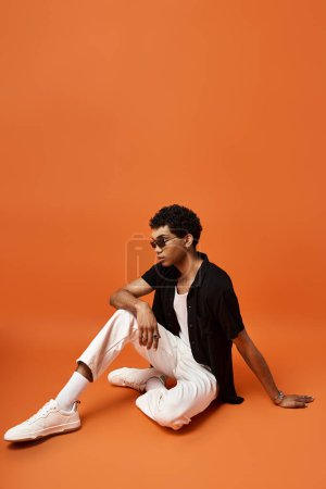 Foto de Hombre afroamericano con gafas de sol sentado en el suelo naranja. - Imagen libre de derechos