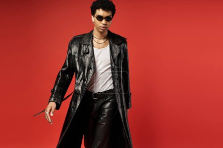 Handsome man in stylish sunglasses, leather trench coat, standing on vibrant red background.