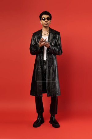 A stylish African American man in a black trench coat standing against a vivid red background.