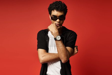 Joven afroamericano con gafas de sol posando con un reloj sobre fondo rojo.