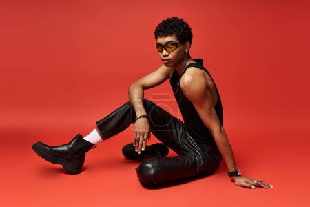Photo for Handsome african american man in leather pants sitting on a red background with stylish sunglasses. - Royalty Free Image