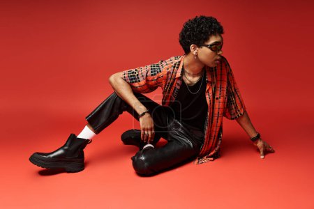A stylish African American man in a red shirt and leather pants sits on the floor.
