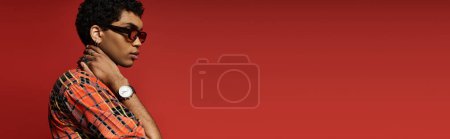 Téléchargez les photos : Bel homme afro-américain frappant une pose dans des lunettes de soleil élégantes sur un fond rouge audacieux. - en image libre de droit
