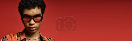 Téléchargez les photos : Beau Afro-Américain avec des lunettes de soleil élégantes debout en toute confiance sur fond rouge. - en image libre de droit