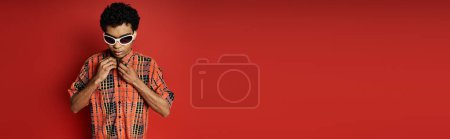 Téléchargez les photos : Beau Afro-Américain en lunettes de soleil pose contre un mur rouge vibrant. - en image libre de droit