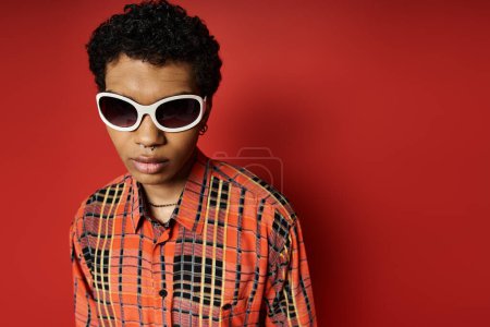 Foto de Hombre afroamericano guapo en gafas de sol de pie frente a una pared roja brillante. - Imagen libre de derechos